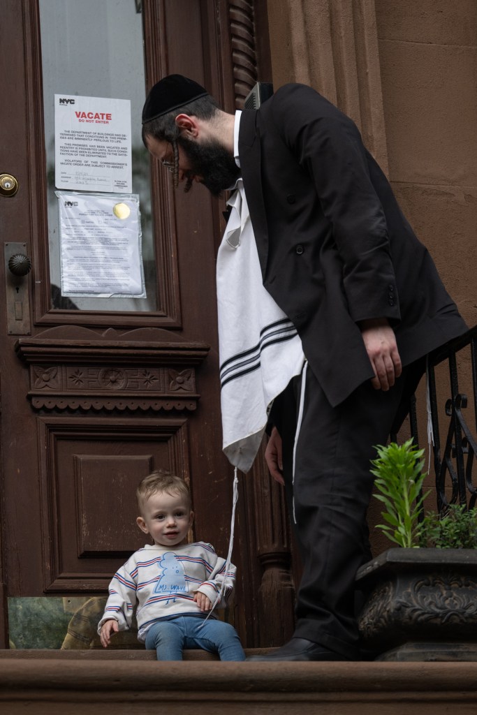 An Orthodox Jewish man with a yarmulke, Jewish prayer shawl and black suit bends over a child in front of a brownstone.