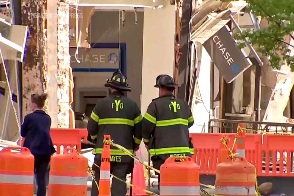 Firefighters standing in front of a partially collapsed 13-story building, with a Chase Bank branch at ground level, following a massive explosion