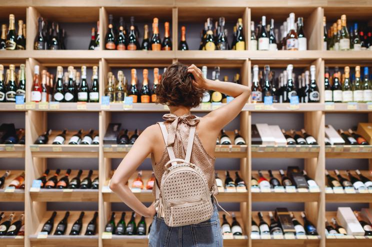Young woman in casual clothes puzzled while choosing a wine bottle in a supermarket