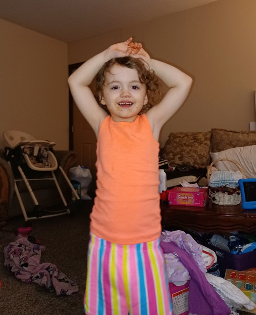 4-year-old Karmity Hoeb seen in an orange tank top and striped colorful pants, smiling for camera with her hands up over her head.