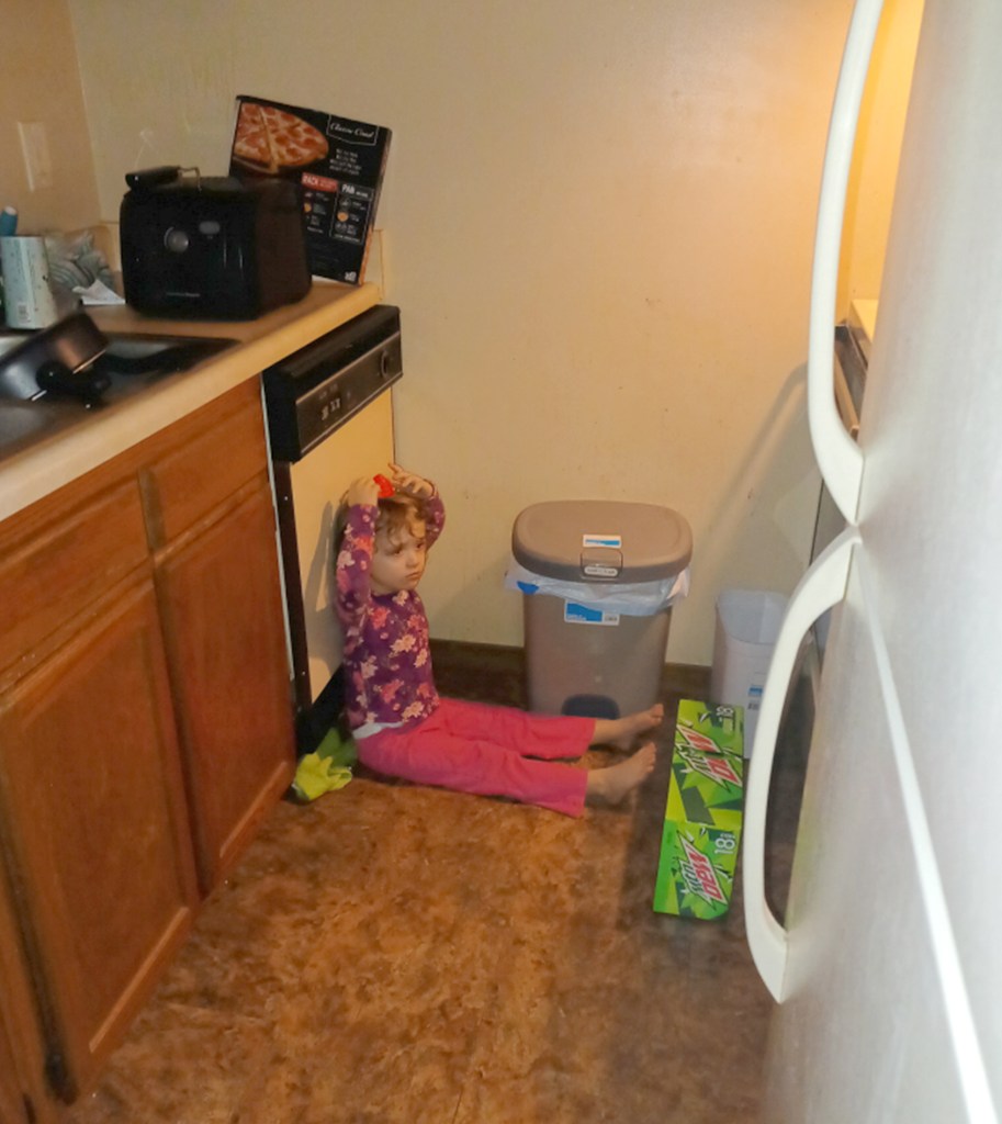 Karmity Hoeb sitting on a kitchen floor next to a garbage pail, with case of mountain dew soda cans at her feet.