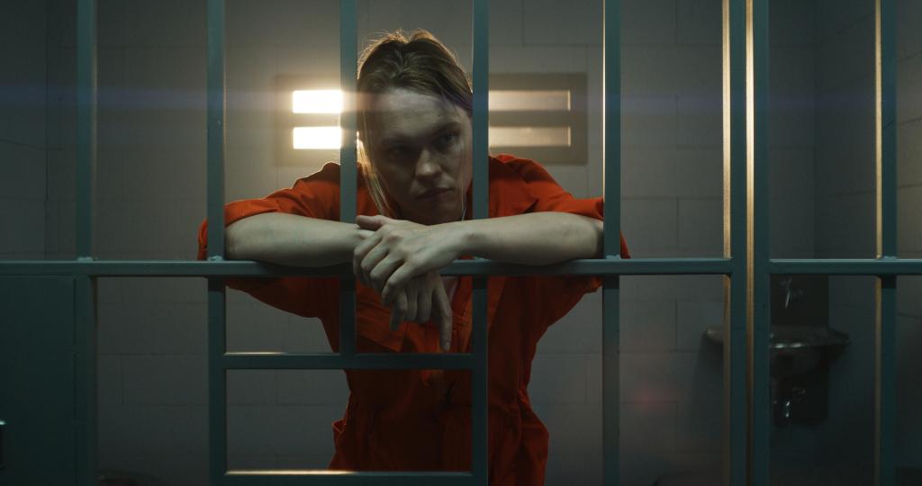 Female prisoner in orange uniform holds hands on metal bars.