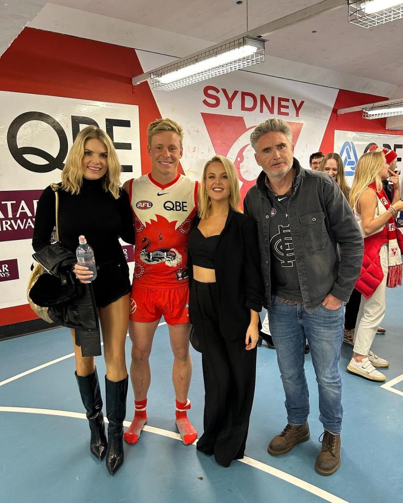 Jana Hocking posing at the center with Dave Hughes on the right and Erin Molan on the left, along with other attendees at a Sydney Swans football match.