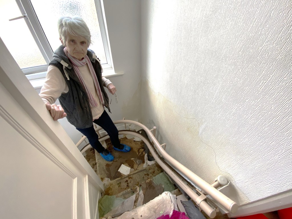 Old woman, Hazel Speed, standing on a staircase in her waterlogged, bug-infested home in Bexley, southeast London.