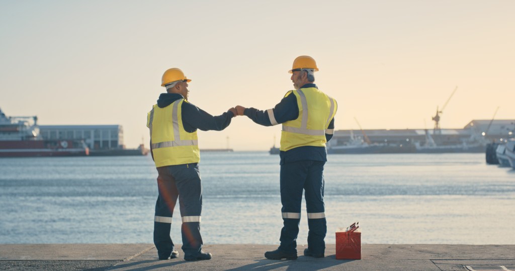 Two logistics, delivery and shipping engineers wearing reflective vests and helmets, giving a fist bump at a cargo dock