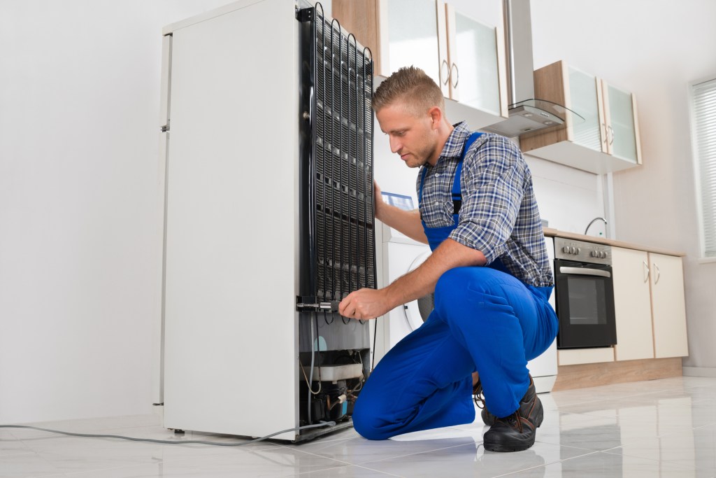 Man in blue overalls repairing a refrigerator with a screwdriver in a house