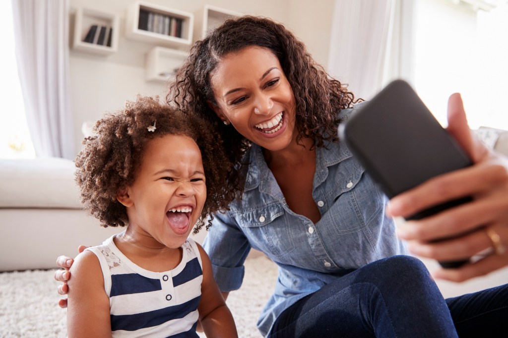 Young mother and toddler daughter laughing while taking a selfie at home