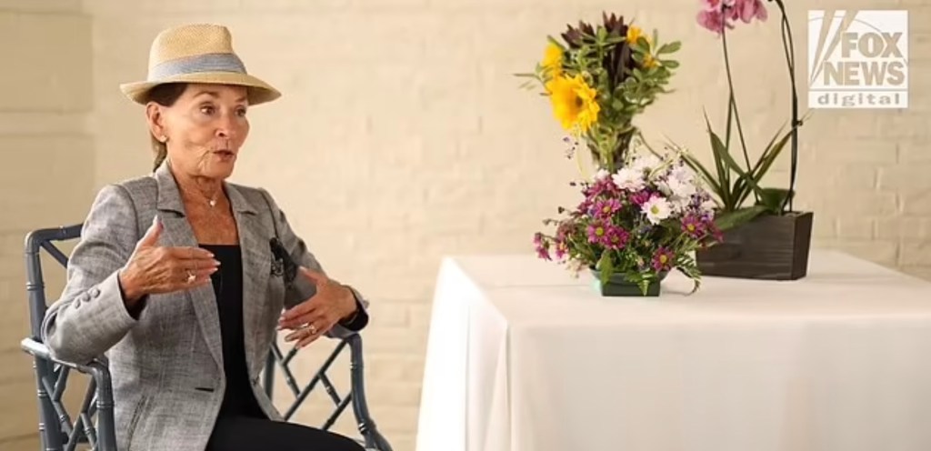 Judith Sheindlin, also known as Judge Judy, sitting in a chair with a hat and flowers on a table, discussing her thoughts on liberal district attorneys in America