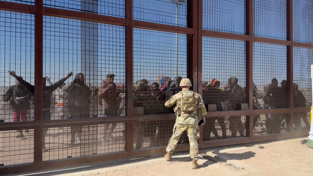 Migrants rush the border wall as guards look on from the US side.
