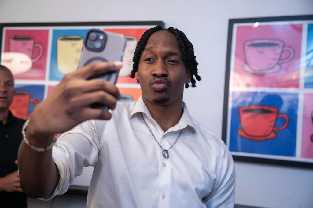 Nate Myles of Harlem, in a white button down shirt with a ring on a silver necklace around his neck, wearing a beaded bracelet and an earring, snapping a selfie at Moka Matcha Cafe, with colorful are of different coffee cups on the wall behind him