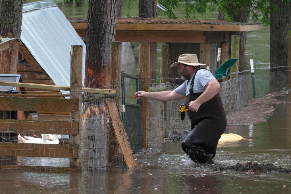 Man in overalls and hat wading through floodwaters in his backyard, preparing his chicken coop amidst severe weather in Liberty, Texas.