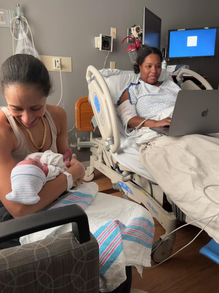 Tamiah Brevard-Rodriguez on her laptop in bed at the hospital while her wife Alyza holds and tends to their newborn boy, Enzo