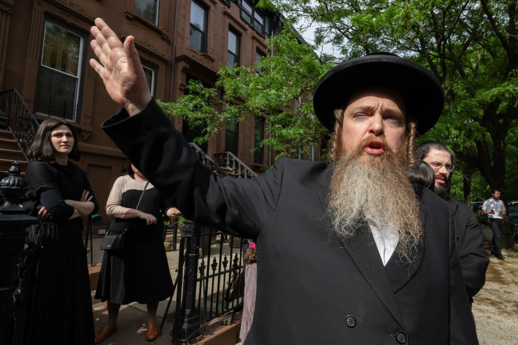 An Orthodox Jew in a black hat, black coat with a long beard and payot talks with his hand up as a couple of women and a man look on.