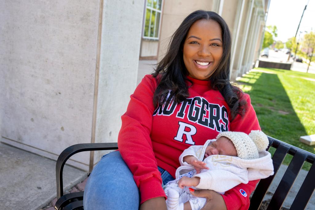 Tamiah Brevard-Rodriguez posing with her new baby boy Enzo while wearing a Rutgers University sweatshirt, sitting on a bench outside