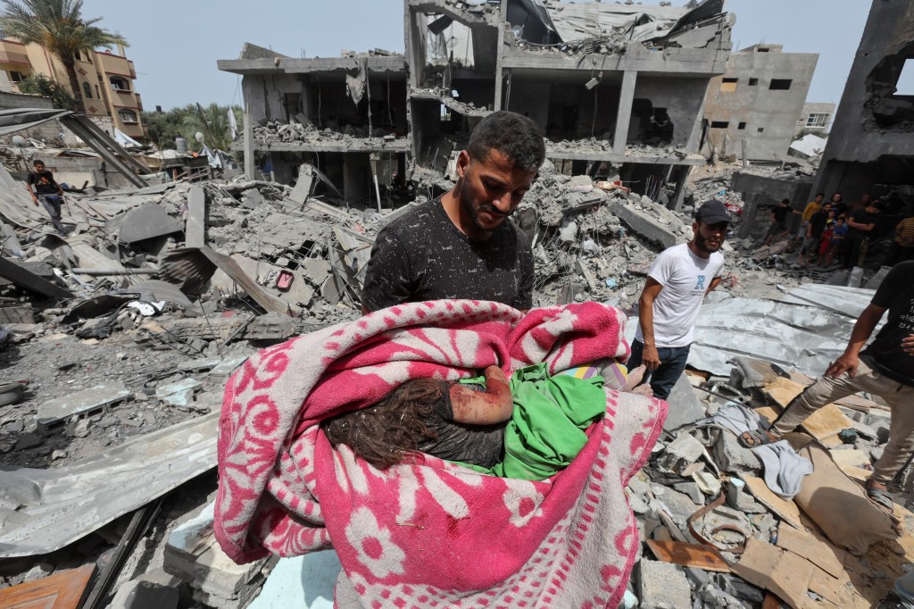 A man standing in building rubble holds the body of a small child in a blanket.