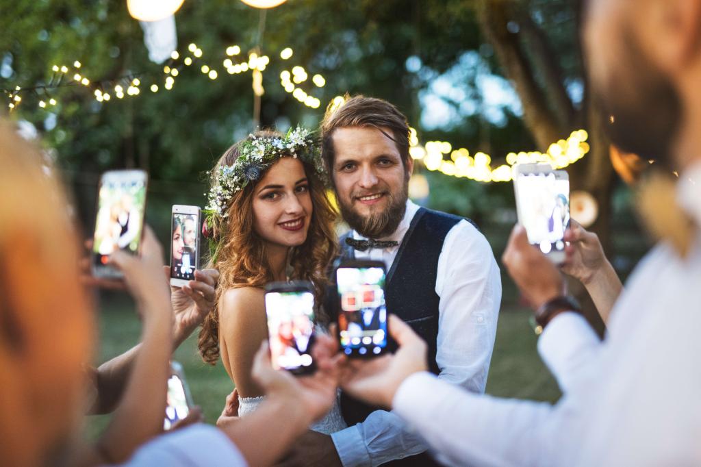 Guests using their smartphones to capture a photo of the bride and groom at an outdoor wedding reception