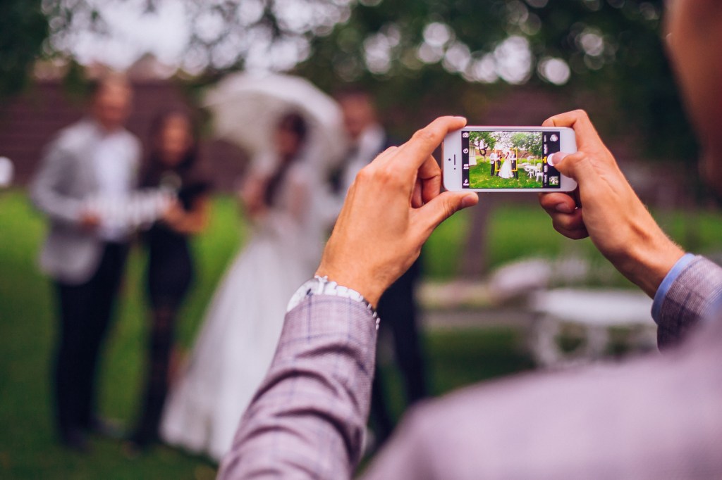 Man holding a smartphone taking a photo of a stylish wedding bride and groom