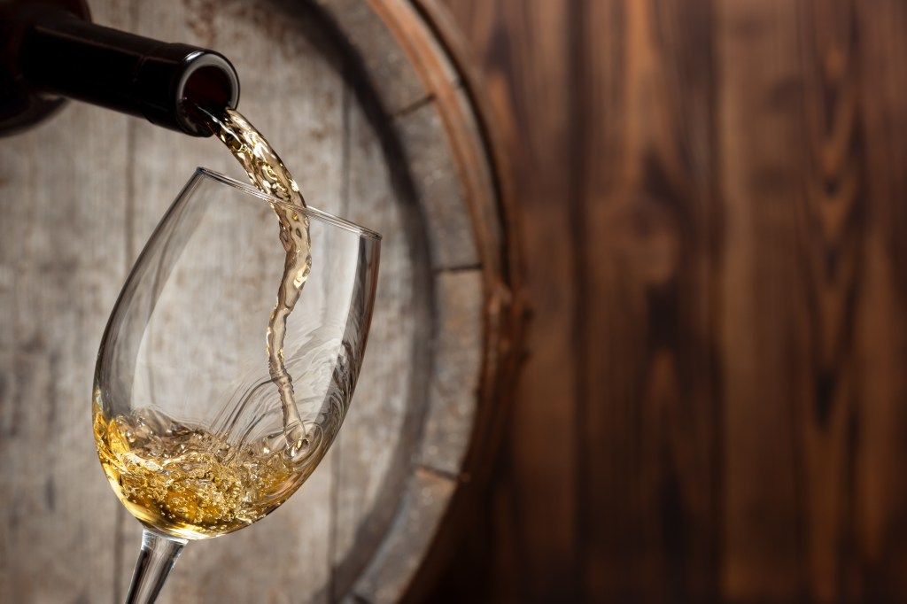 White wine being poured from a bottle into a glass with an old wooden barrel in the background