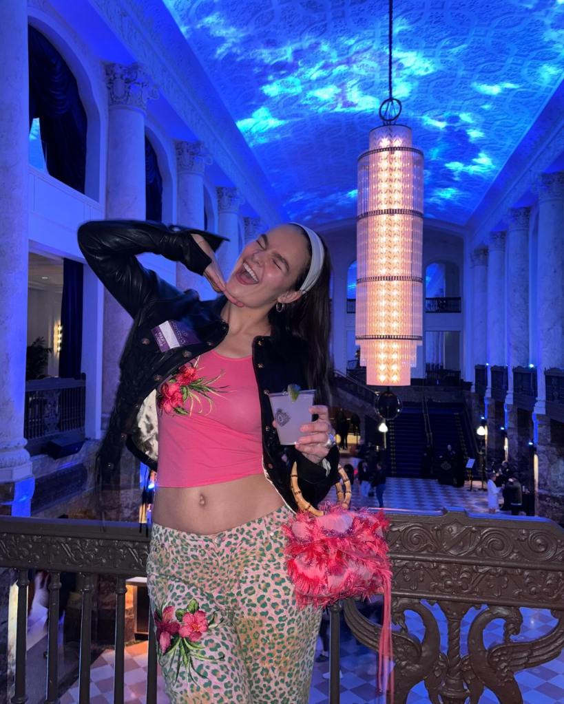 A woman in a midriff holds a drink and smiles widely in front of a chandelier at the Brooklyn Paramount Historic Theater. 