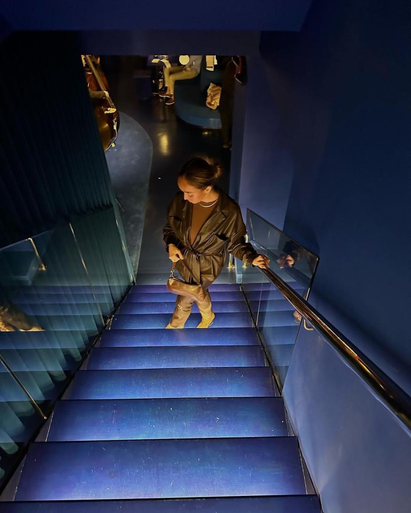 A woman wears a leather trench and poses looking down on the blue stairs at Only Love Strangers. 