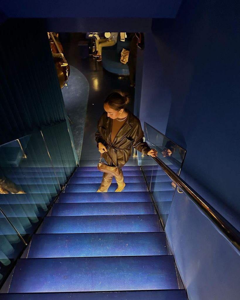 A woman wears a leather trench and poses looking down on the blue stairs at Only Love Strangers. 