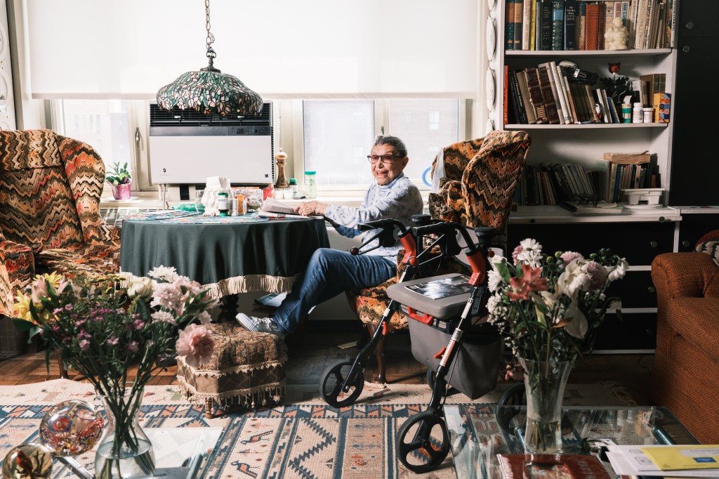 Interior Michelle Cohen in her apartment. Wide shot shows table, chairs, flowers on table, walker. 