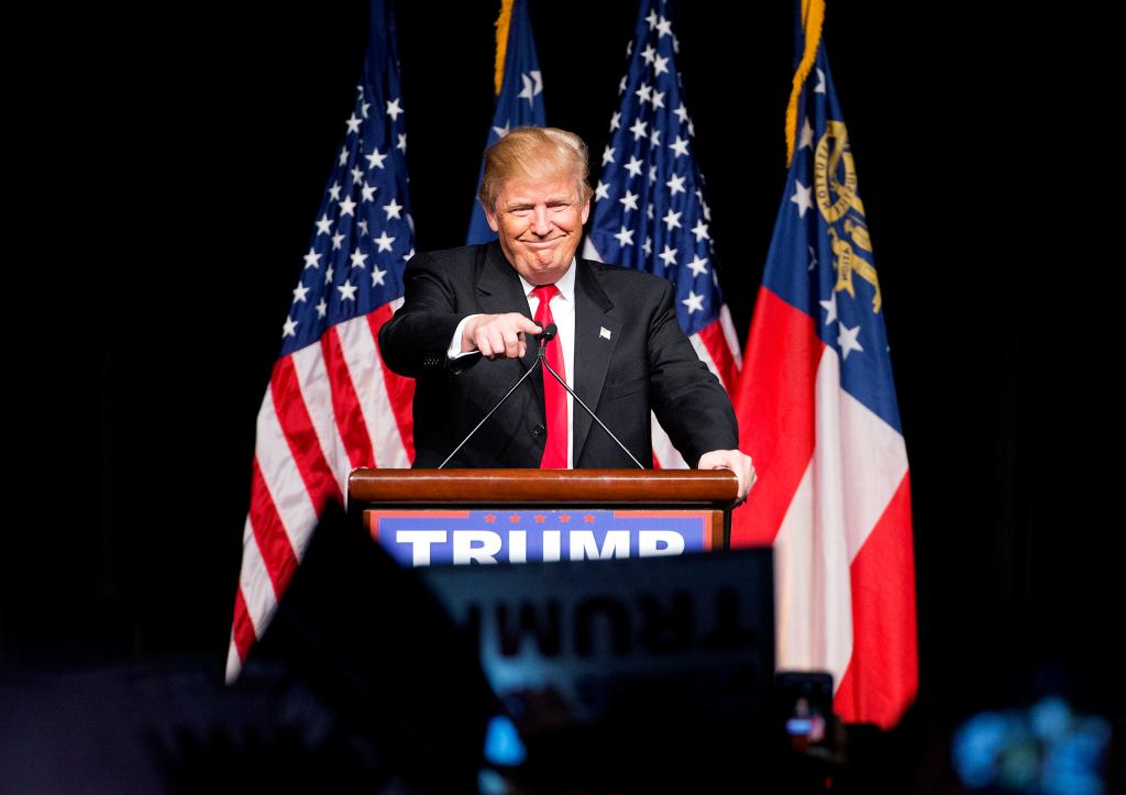 Donald Trump pointing to the crowd while standing at a podium with flags behind him at a 2016 campaign event in Atlanta