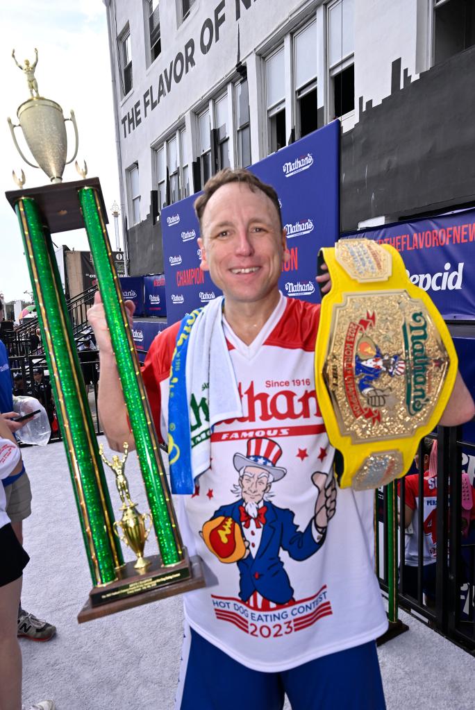 Joey 'JAWS' Chestnut holding a trophy and a belt after winning the 2023 Nathan's Fourth of July Hot Dog contest, having eaten 62 hot dogs in 10 minutes