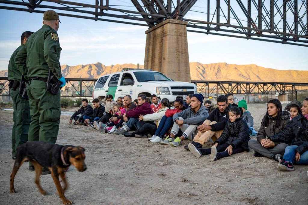 A couple dozen migrants sit on the ground as two Border Patrol agents guard them.