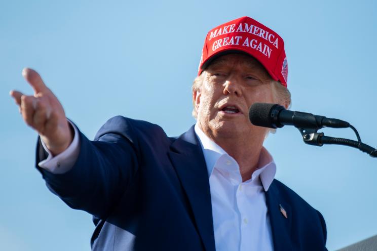 Former President Donald Trump speaking from a podium at a rally in Greenwood, Nebraska, while wearing a red hat and pointing at a microphone