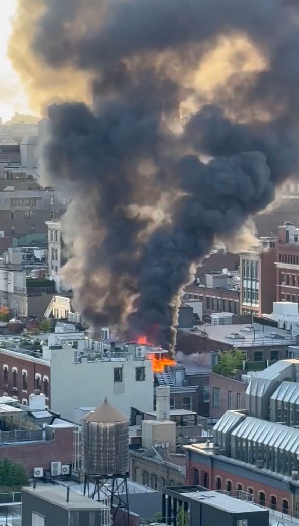 Man on neighboring rooftop using a garden hose to help extinguish a fire on the roof of 463 Broome Street in SoHo, Manhattan, with black smoke billowing from the building
