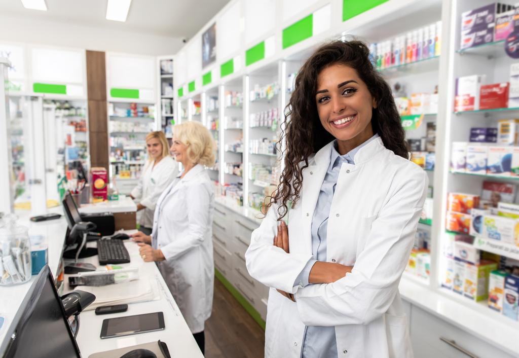 A female pharmacist in a white coat standing in front of a counter with other people in the background
