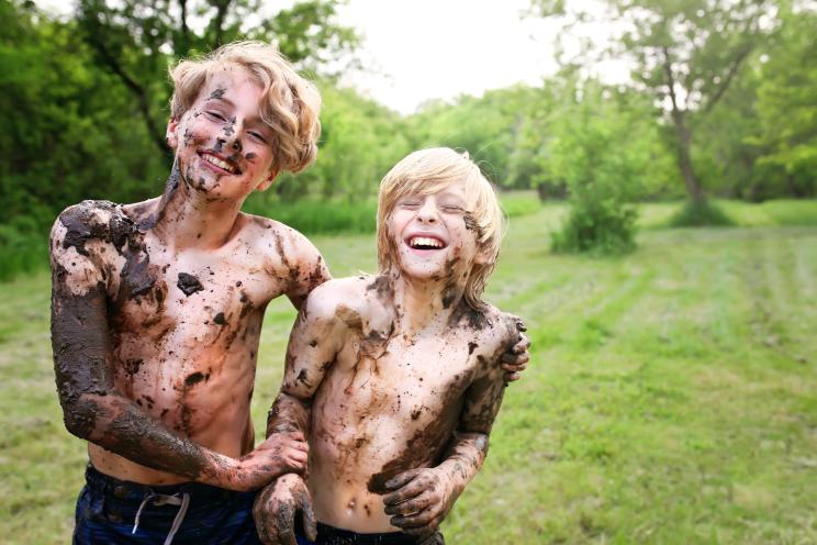 Two joyous children covered in mud laughing near a river on a summer day