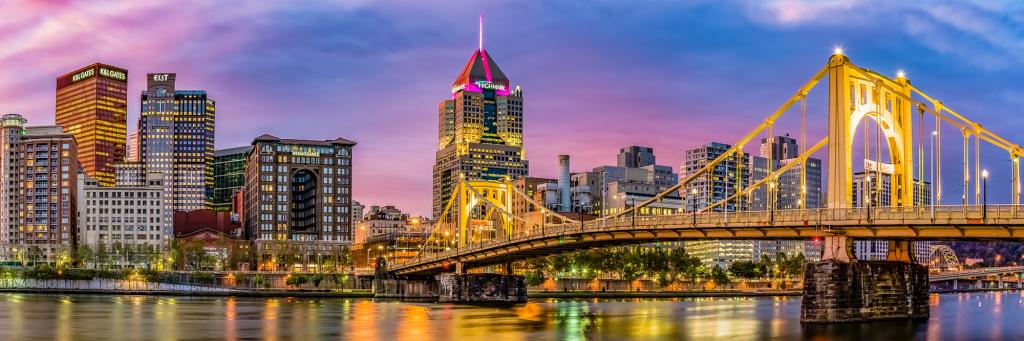 Panoramic view of Pittsburgh cityscape from North Shore, featuring a bridge over a river and buildings in the background at dawn