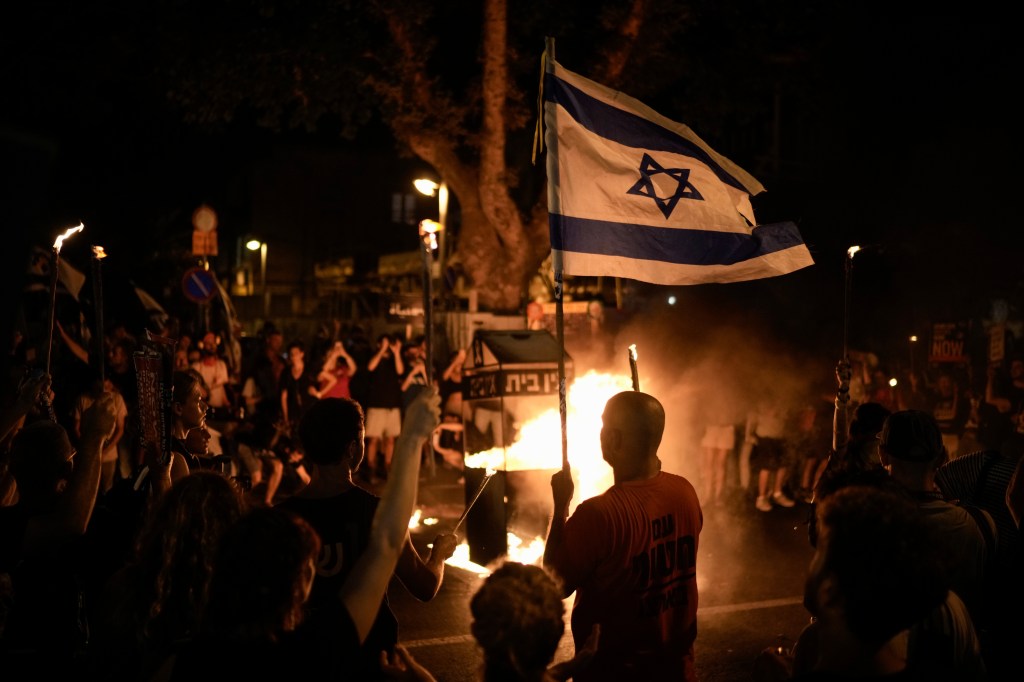 Protesters in Tel Aviv, Israel holding a flag and standing in front of a fire, protesting against Israeli Prime Minister Benjamin Netanyahu's government and demanding release of Gaza Strip hostages