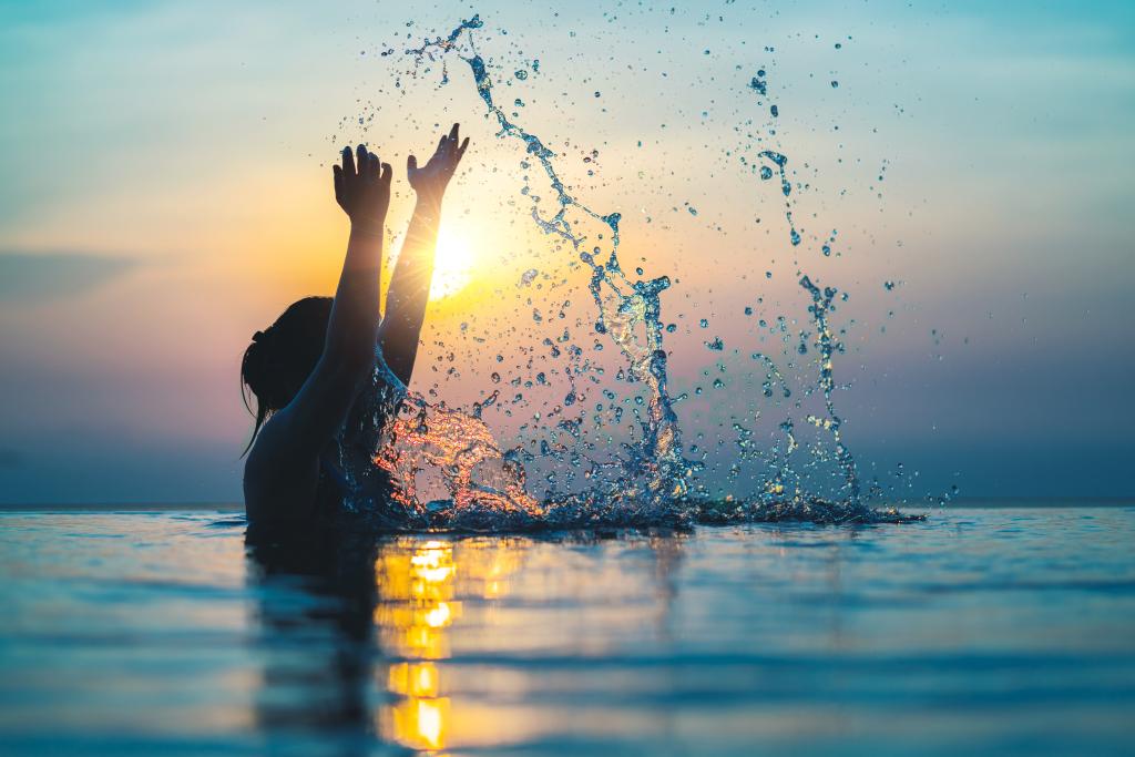 Black silhouette of asian woman splash water on summer vacation holiday relaxing in infinity swimming pool with blue sea sunset view. Healthy happiness lifestyle