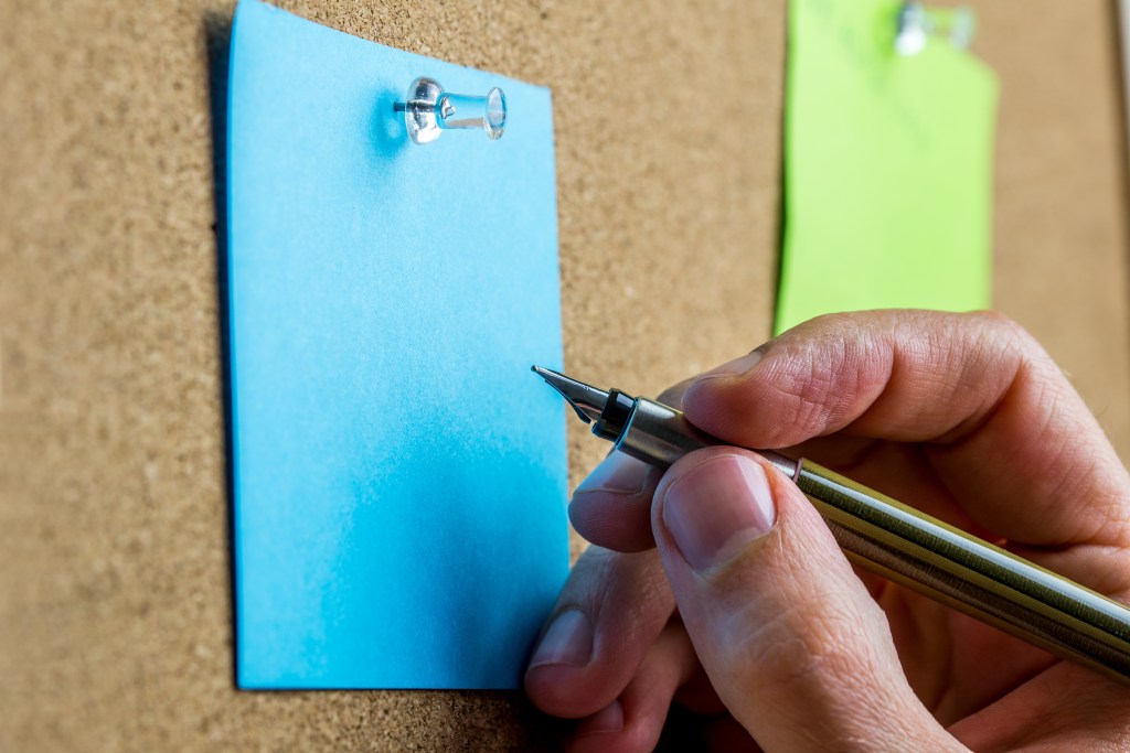 Male hand writing on blank sheet of blue paper with copyspace pinned up on a cork bulletin board.