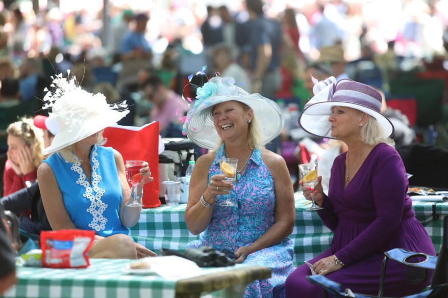 Three woman wearing hats sit together as they drink cocktails at a pic nic table