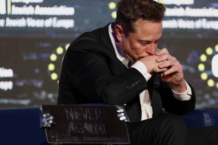 Tesla CEO Elon Musk wears a black suit as he sits with his mouth on his hands.