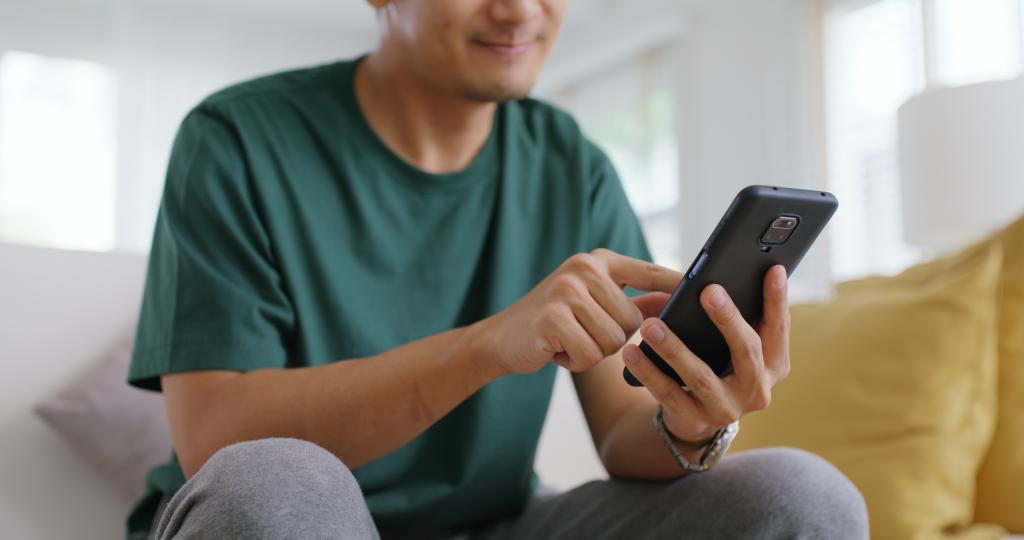 Single Asian man sitting on sofa at home, smiling while looking at a girl's photo on a dating app on his phone