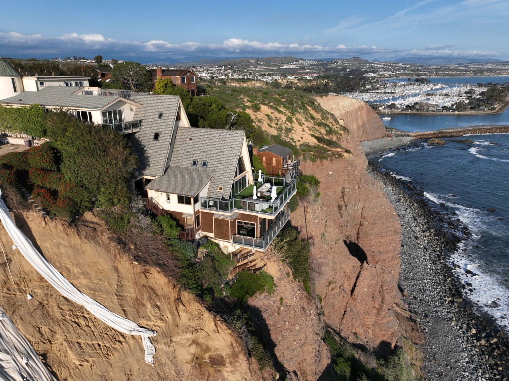 An aerial view of a double A-frame cliff-side mansion, between two other mansions affected by a landslide and protective tarps to protect from further slides from upcoming storms in Dana Point Tuesday, Feb. 27, 2024. 