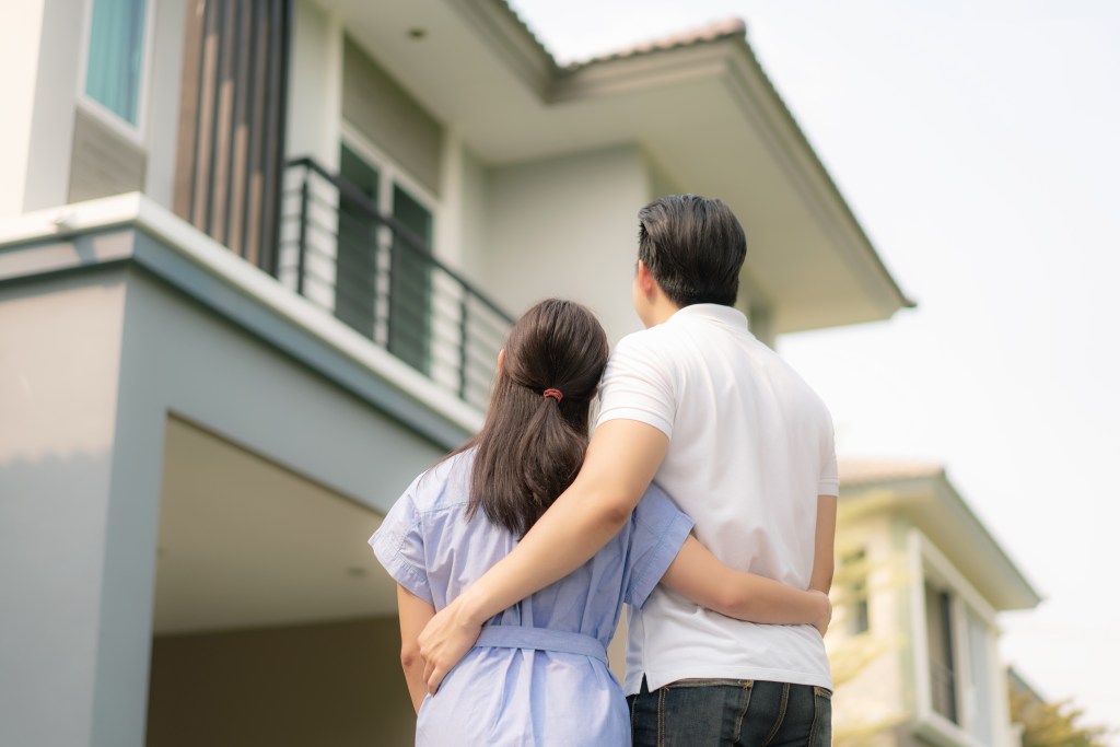 Back portrait of Asian young couple standing and hugging together looking happy in front of their new house to start new life