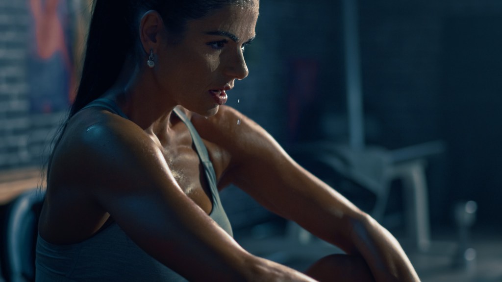 Fit brunette woman in sportswear, catching her breath in an industrial gym after intense workout, motivational posters in background.