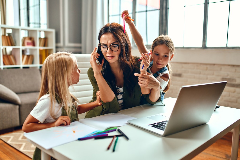 Business woman working on laptop while her two daughters play and interfere at home table, emphasizing work-life balance