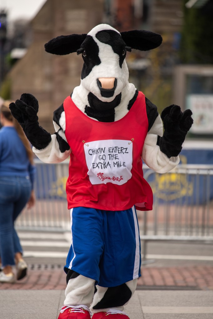 A cow mascot wearing running shorts as well as a race bib with "chikin eaterz go the extra mile" pinned to a running jersey, strikes a pose on April 16, 2022 in Copley Square in Boston, MA