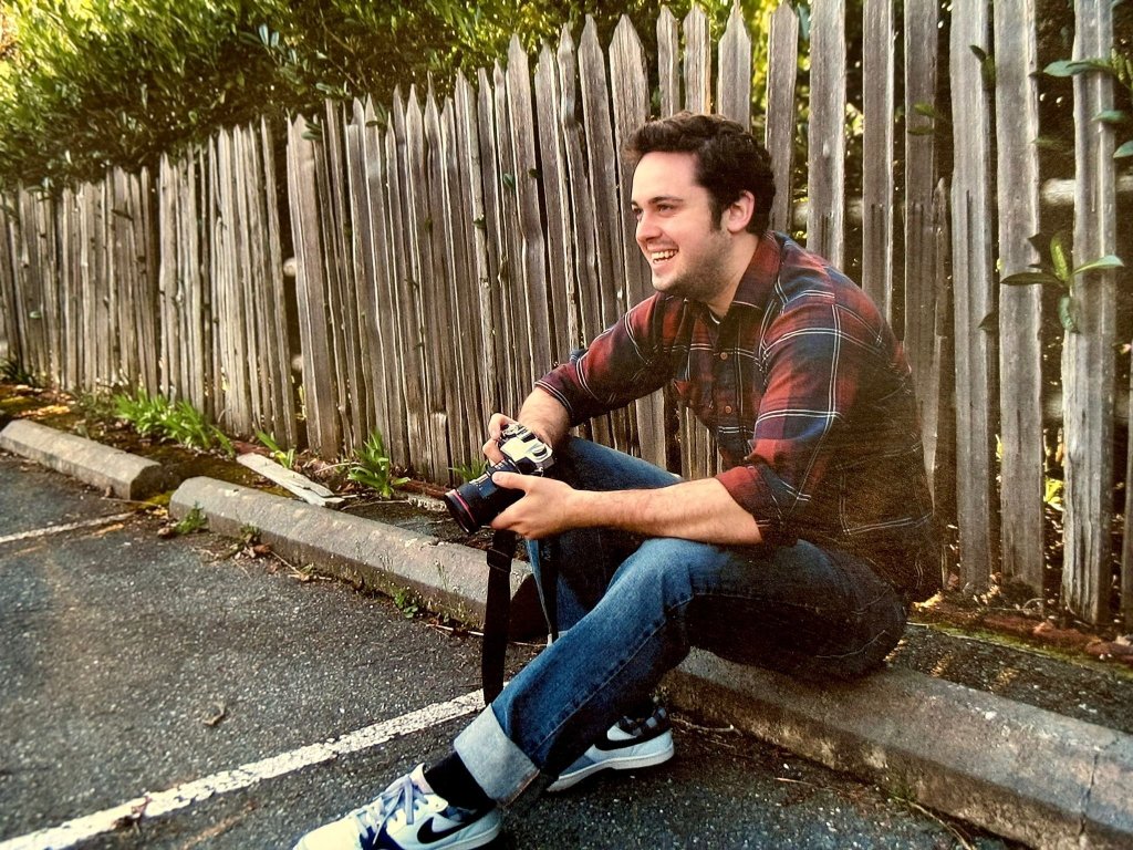 A man, Brandon Young, sitting on a curb holding a camera at a 'FUNeral' event with a bouncy castle and party bags in the background.