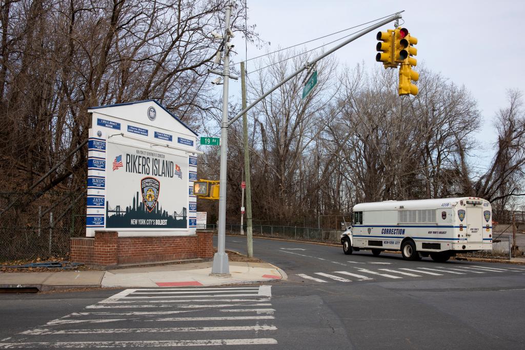 A Department of Correction bus driving up to the entrance of the bridge that connects to Rikers Island near a sign that says Rikers Island