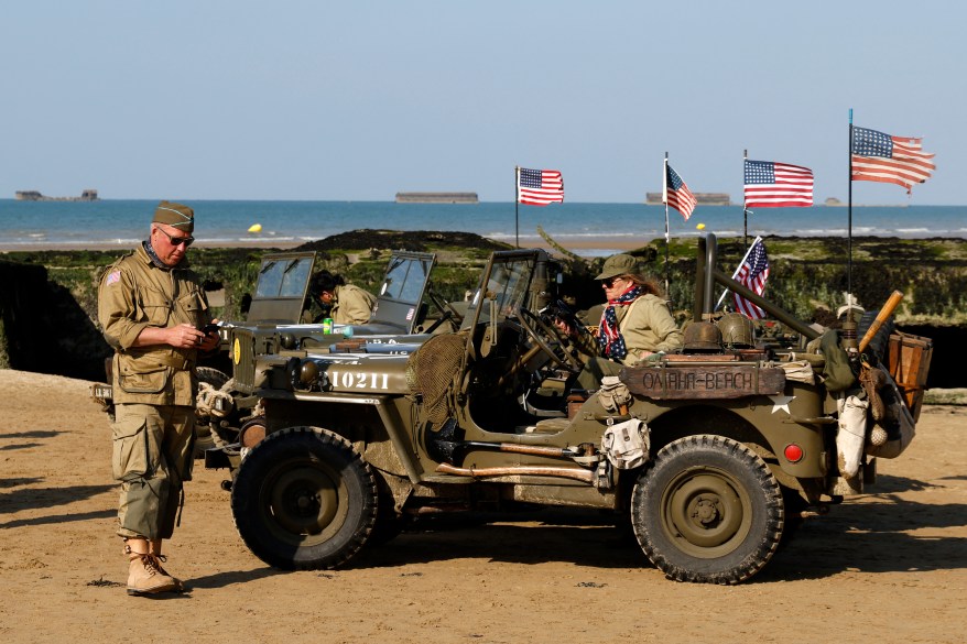 Enthusiasts dressed in replica WWII-era military attire park Willys Jeeps on the beach, as Phoenix breakwaters forming part of the Mulberry harbour are seen out at sea on the horizon, at Arromanches-les-Bains, northwestern France, on June 5, 2024.