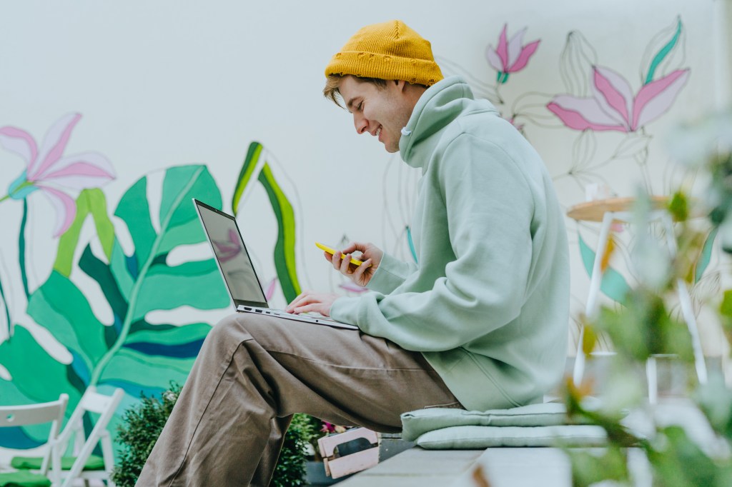 Young freelancer man using laptop and smartphone while sitting on outdoor stairs