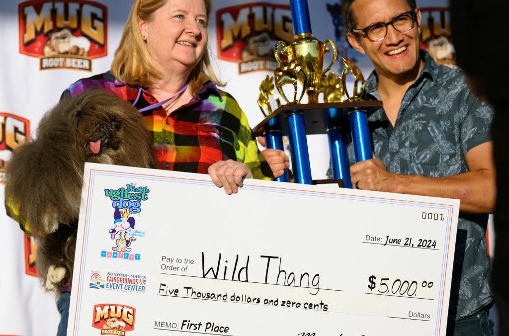 A woman holds a dog in one hand and in the other, an oversized check made out to "Wild Thang" for $5,000, while a man holds a big trophy behind her.